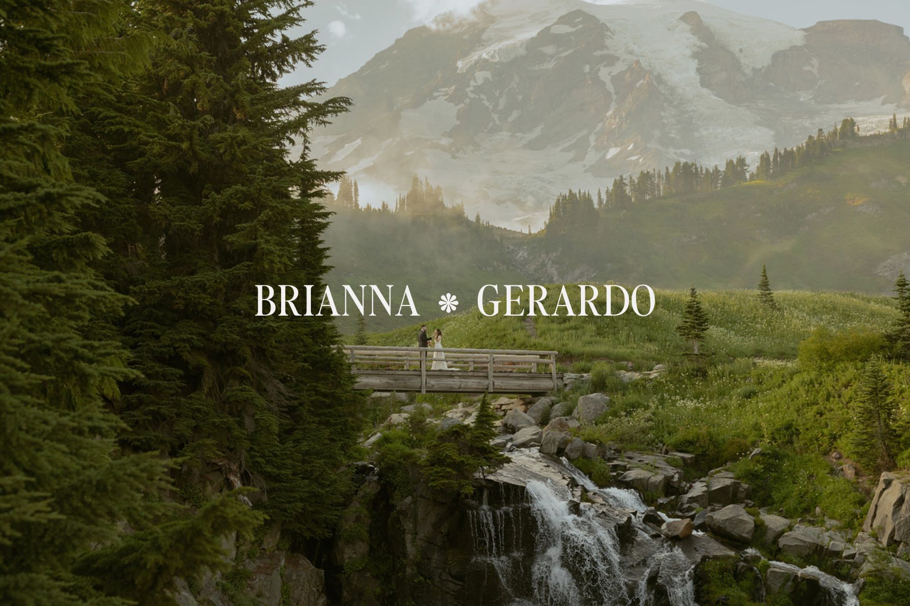 Couple standing on a wooden bridge during their Mount Rainier elopement, surrounded by wildflowers, evergreens, and a waterfall with the mountain glowing behind them.